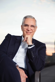 An individual with short gray hair and glasses is seated, wearing a formal dark suit and a white shirt. Their posture is relaxed with one hand resting on their chin, conveying a sense of confidence and ease. The background appears to be an outdoor setting with a blurred view of a cityscape at dusk, adding a serene atmosphere to the image.
