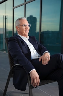 A mature man with grey hair wearing glasses, a white shirt, and a dark suit, sitting and smiling outdoors against a backdrop of large glass windows reflecting an urban cityscape.