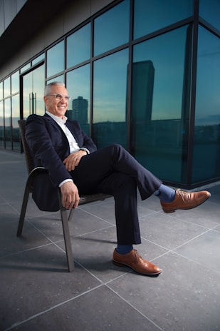 A man in a suit sits confidently on a chair with his leg crossed, located on a modern terrace with large glass windows reflecting the cityscape. He is smiling and appears relaxed, dressed in a dark suit with brown leather shoes and blue socks.