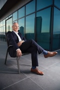 A man in a suit sits confidently on a chair with his leg crossed, located on a modern terrace with large glass windows reflecting the cityscape. He is smiling and appears relaxed, dressed in a dark suit with brown leather shoes and blue socks.