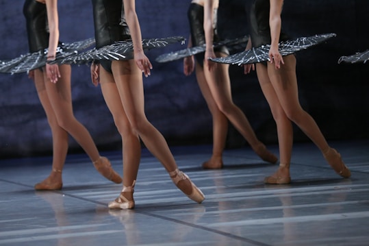 Ballet dancers in tutus are poised on stage, demonstrating graceful movements and coordination. The focus is on their legs, showing strength and elegance, as they balance on pointe shoes.