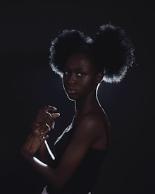 A woman with an afro hairstyle is illuminated from the back, creating a rim of light around her silhouette. Her pose is graceful and elegant, with hands gently intertwined and a contemplative expression. The lighting is dramatic, enhancing the textures and contours of her hair and skin.