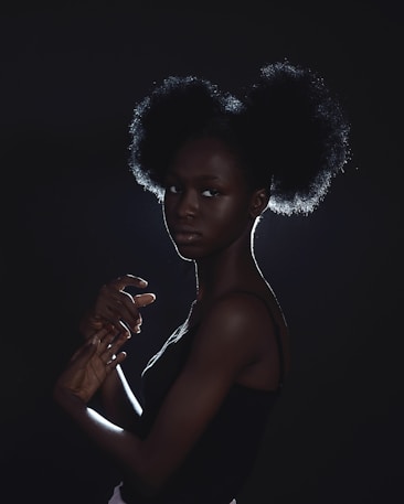 A woman with an afro hairstyle is illuminated from the back, creating a rim of light around her silhouette. Her pose is graceful and elegant, with hands gently intertwined and a contemplative expression. The lighting is dramatic, enhancing the textures and contours of her hair and skin.