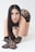 A woman poses on her hands and knees, wearing black lace gloves and a matching lace outfit. She has long dark hair and a neutral expression. The background is plain and light-colored.