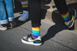 Five legs are visible, with four people wearing colorful rainbow-striped socks. The individuals are standing on a sunlit sidewalk, casting shadows. Some are wearing casual sneakers, with one pair of shoes having a floral design.