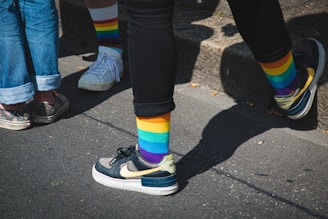 Five legs are visible, with four people wearing colorful rainbow-striped socks. The individuals are standing on a sunlit sidewalk, casting shadows. Some are wearing casual sneakers, with one pair of shoes having a floral design.