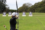 An individual is practicing archery in an open field with multiple targets set at varying distances. The person has a bow drawn back, aiming at the targets which feature traditional bullseye patterns. Surrounding the field are trees, creating a natural setting.