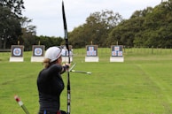 An individual is practicing archery in an open field with multiple targets set at varying distances. The person has a bow drawn back, aiming at the targets which feature traditional bullseye patterns. Surrounding the field are trees, creating a natural setting.