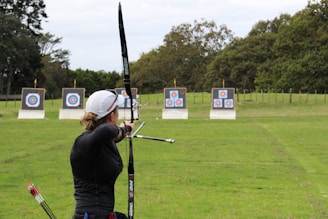 An individual is practicing archery in an open field with multiple targets set at varying distances. The person has a bow drawn back, aiming at the targets which feature traditional bullseye patterns. Surrounding the field are trees, creating a natural setting.