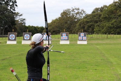 An individual is practicing archery in an open field with multiple targets set at varying distances. The person has a bow drawn back, aiming at the targets which feature traditional bullseye patterns. Surrounding the field are trees, creating a natural setting.