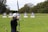 An individual is practicing archery in an open field with multiple targets set at varying distances. The person has a bow drawn back, aiming at the targets which feature traditional bullseye patterns. Surrounding the field are trees, creating a natural setting.