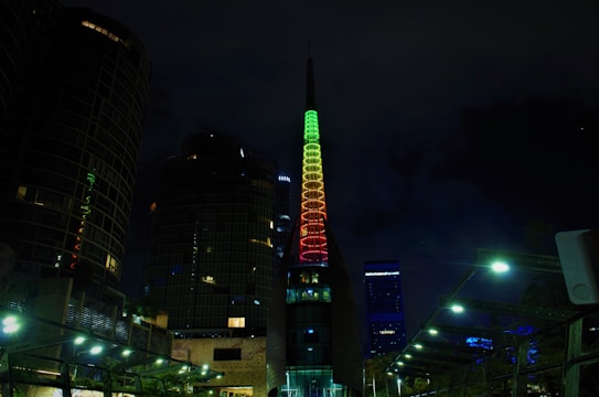 The image depicts a night-time cityscape featuring a tall, modern building with a spire illuminated in a gradient of green, yellow, and red colors. Surrounding the building are other high-rise structures, and the scene is lit by streetlights providing a soft glow to the area.