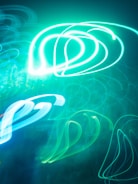 Bright and swirling neon lights create abstract patterns against a dark background, with predominant hues of teal and blue forming fluid shapes.
