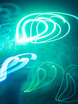 Bright and swirling neon lights create abstract patterns against a dark background, with predominant hues of teal and blue forming fluid shapes.