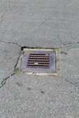 A metal drain cover is set into cracked asphalt pavement. Some small patches of grass are visible around the edges of the drain.