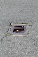 A metal drain cover is set into cracked asphalt pavement. Some small patches of grass are visible around the edges of the drain.