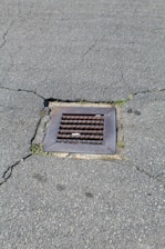 A metal drain cover is set into cracked asphalt pavement. Some small patches of grass are visible around the edges of the drain.