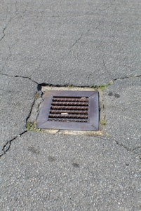 A metal drain cover is set into cracked asphalt pavement. Some small patches of grass are visible around the edges of the drain.
