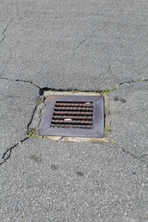 A metal drain cover is set into cracked asphalt pavement. Some small patches of grass are visible around the edges of the drain.
