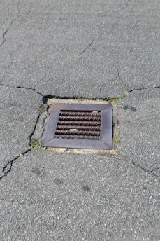 A metal drain cover is set into cracked asphalt pavement. Some small patches of grass are visible around the edges of the drain.