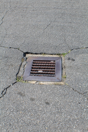 A metal drain cover is set into cracked asphalt pavement. Some small patches of grass are visible around the edges of the drain.