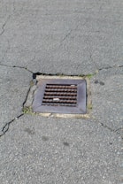 A metal drain cover is set into cracked asphalt pavement. Some small patches of grass are visible around the edges of the drain.
