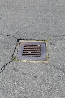 A metal drain cover is set into cracked asphalt pavement. Some small patches of grass are visible around the edges of the drain.