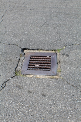 A metal drain cover is set into cracked asphalt pavement. Some small patches of grass are visible around the edges of the drain.