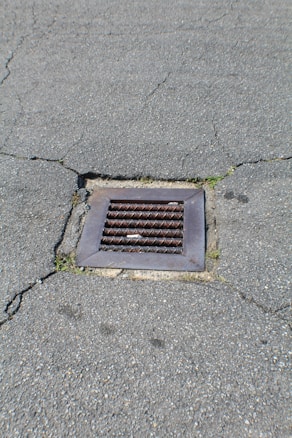 A metal drain cover is set into cracked asphalt pavement. Some small patches of grass are visible around the edges of the drain.