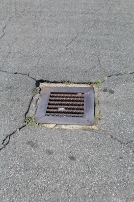 A metal drain cover is set into cracked asphalt pavement. Some small patches of grass are visible around the edges of the drain.
