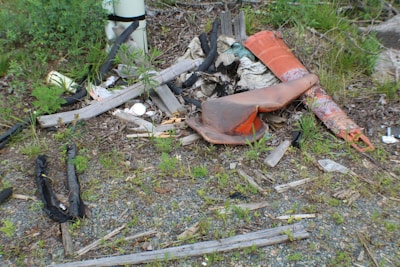 A cluttered scene with various debris including wooden planks, an old orange traffic cone, and plastic materials scattered on the ground amidst patches of green grass and plants.