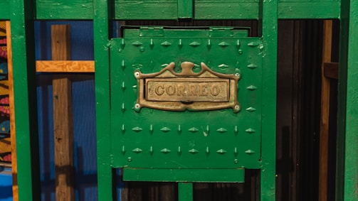 A green metal mailbox with an ornate brass plate displaying the word 'Correo'. The mailbox is set against a background that includes wooden elements and a blue panel, contributing to a colorful and rustic appearance.