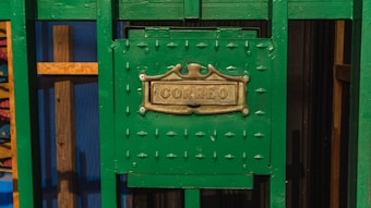 A green metal mailbox with an ornate brass plate displaying the word 'Correo'. The mailbox is set against a background that includes wooden elements and a blue panel, contributing to a colorful and rustic appearance.