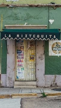 A small storefront with a weathered green facade and a dark green awning displaying the text 'La Mixtequita'. The door is covered with various colorful posters and promotional flyers. The building shows signs of wear, with peeling paint and noticeable cracks.