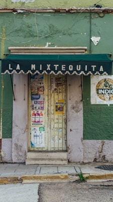 A small storefront with a weathered green facade and a dark green awning displaying the text 'La Mixtequita'. The door is covered with various colorful posters and promotional flyers. The building shows signs of wear, with peeling paint and noticeable cracks.