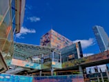 Urban architecture scene with modern buildings featuring reflective surfaces. A tall structure with a colorful, mosaic-like facade stands prominently against a bright blue sky. Nearby, other buildings have iridescent, shiny panels and glass canopies. People are seen walking on an elevated walkway.