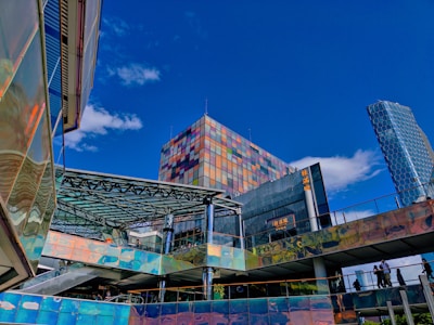 Urban architecture scene with modern buildings featuring reflective surfaces. A tall structure with a colorful, mosaic-like facade stands prominently against a bright blue sky. Nearby, other buildings have iridescent, shiny panels and glass canopies. People are seen walking on an elevated walkway.