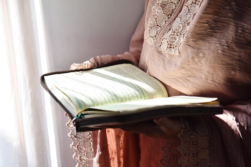 A person in a robe with lace details holds an open book. Sunlight illuminates the pages, revealing text written in a non-Latin script.