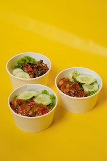 Three round, white disposable bowls filled with a colorful mix of diced vegetables and garnished with fresh cucumber slices and leafy greens are arranged on a solid bright yellow background.