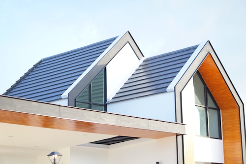 A modern house with a minimalist design featuring sharp angles and a clean aesthetic. The roof is made of dark gray slate tiles, and the walls are primarily white with wooden accents. Large windows allow natural light to flood the interior. The house has a sleek overhang and a prominent triangular peak.