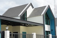 A modern house with a gabled roof design. The exterior features white walls contrasted with a teal accent and large windows. The roof is composed of dark gray tiles, and there is a black and white gate in front. A lamp post is visible at the front of the property, and there is a metal fence surrounding the area.