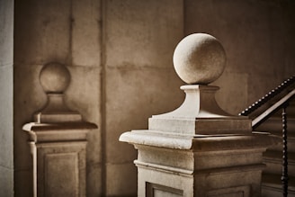 Stone balustrades with spherical finials are positioned in a dimly lit, classical architectural space. The lighting creates soft shadows and highlights the texture of the stone surface. A staircase with ornate railings is partially visible in the background, lending an air of elegance and history to the setting.