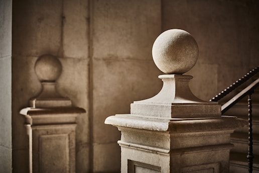 Stone balustrades with spherical finials are positioned in a dimly lit, classical architectural space. The lighting creates soft shadows and highlights the texture of the stone surface. A staircase with ornate railings is partially visible in the background, lending an air of elegance and history to the setting.