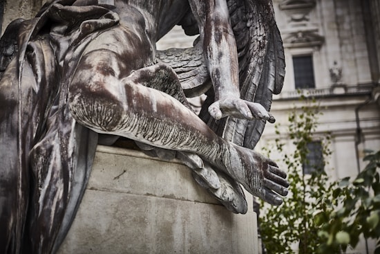 A close-up view of a detailed stone statue depicting an angelic figure with wings. The statue's posture suggests a relaxed, almost reclining pose, with one arm extended and fingers gently resting. The surface of the statue shows signs of weathering, adding a sense of antiquity. The background features a blurred architectural structure, and some greenery is visible.