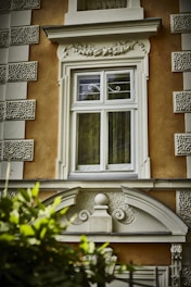 A decorative window frame on an orange building facade features intricate architectural details, including ornate molding and stucco textures. A mixture of classical and baroque design elements enriches the structure. Green leaves partially obscure the bottom of the image.