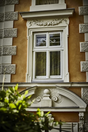 A decorative window frame on an orange building facade features intricate architectural details, including ornate molding and stucco textures. A mixture of classical and baroque design elements enriches the structure. Green leaves partially obscure the bottom of the image.