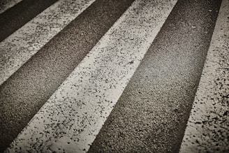 Several worn, white zebra stripes on a rough asphalt road surface. The texture of the asphalt is visible, with cracks and small stones embedded in the pavement.