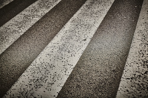 Several worn, white zebra stripes on a rough asphalt road surface. The texture of the asphalt is visible, with cracks and small stones embedded in the pavement.