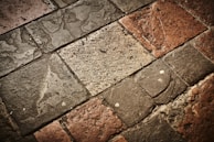 A textured surface composed of irregularly shaped stone tiles in various shades of brown, grey, and beige. The stones have a worn appearance with visible imperfections and some patina.