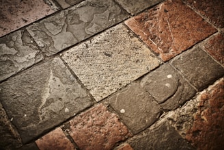 A textured surface composed of irregularly shaped stone tiles in various shades of brown, grey, and beige. The stones have a worn appearance with visible imperfections and some patina.